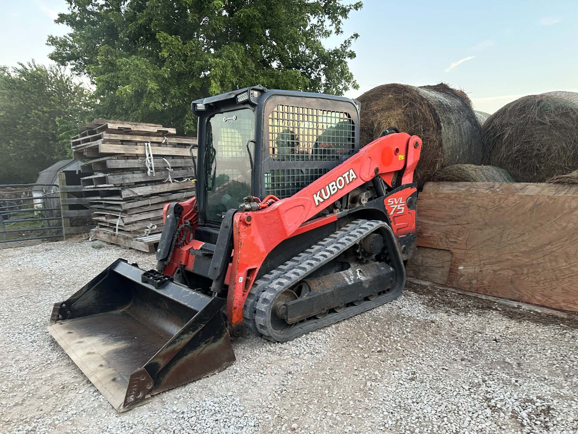 A red Kubota compact track loader with a front bucket parked on a gravel lot next to large round hay bales.