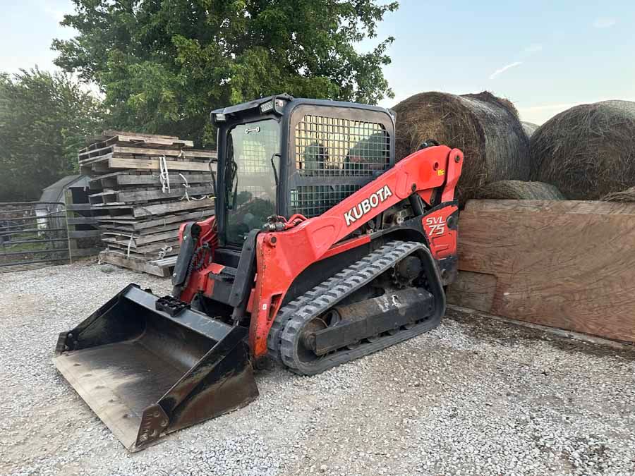 An orange Kubota compact track loader with a front bucket parked on a gravel lot next to stacked hay bales.