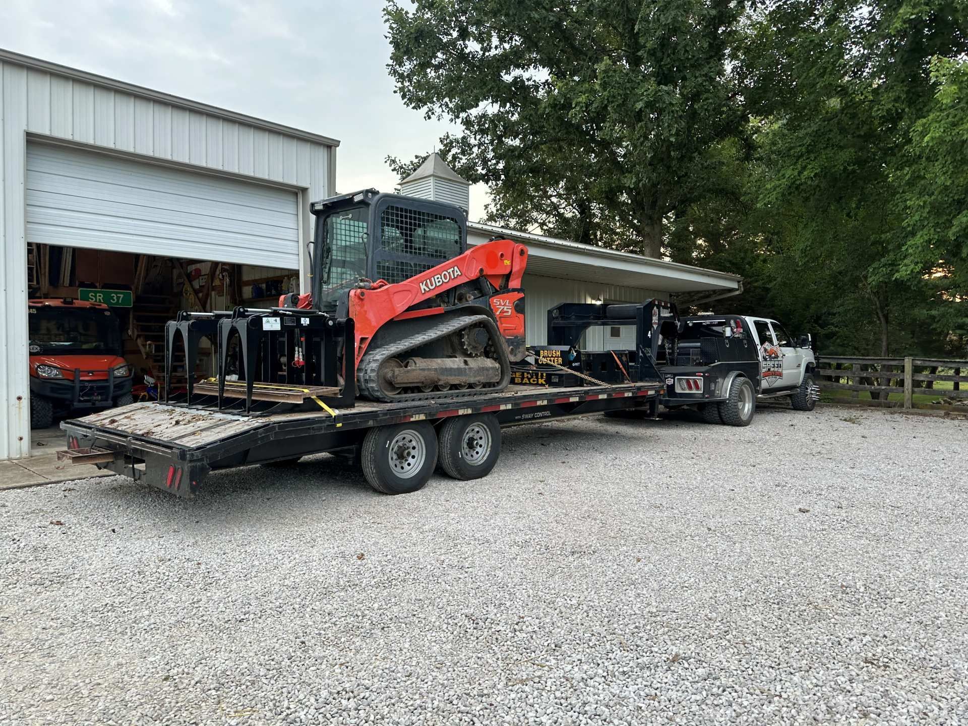 An orange compact track loader sits on a flatbed trailer attached to a white truck on a gravel lot near a storage building.