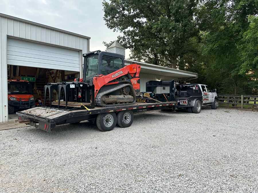 A bright orange skid steer with a grapple attachment sits on a flatbed trailer towed by a truck in a gravel yard.
