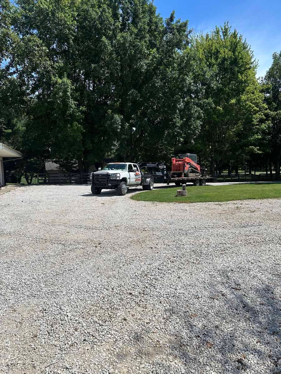 A white pickup truck pulling a trailer with orange construction equipment on a gravel driveway near trees.