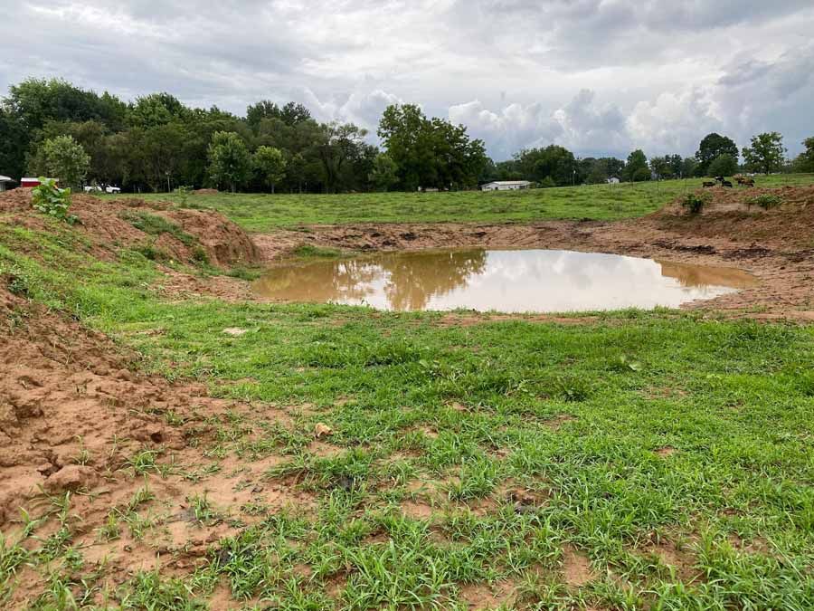 A muddy pond sits in the center of a grassy field with piles of excavated dirt under a cloudy sky.
