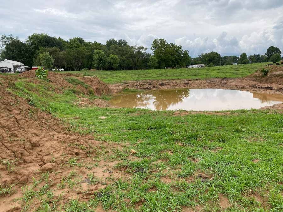 A muddy pond sits in a cleared, grassy field under a cloudy sky, bordered by piles of excavated earth and distant trees.