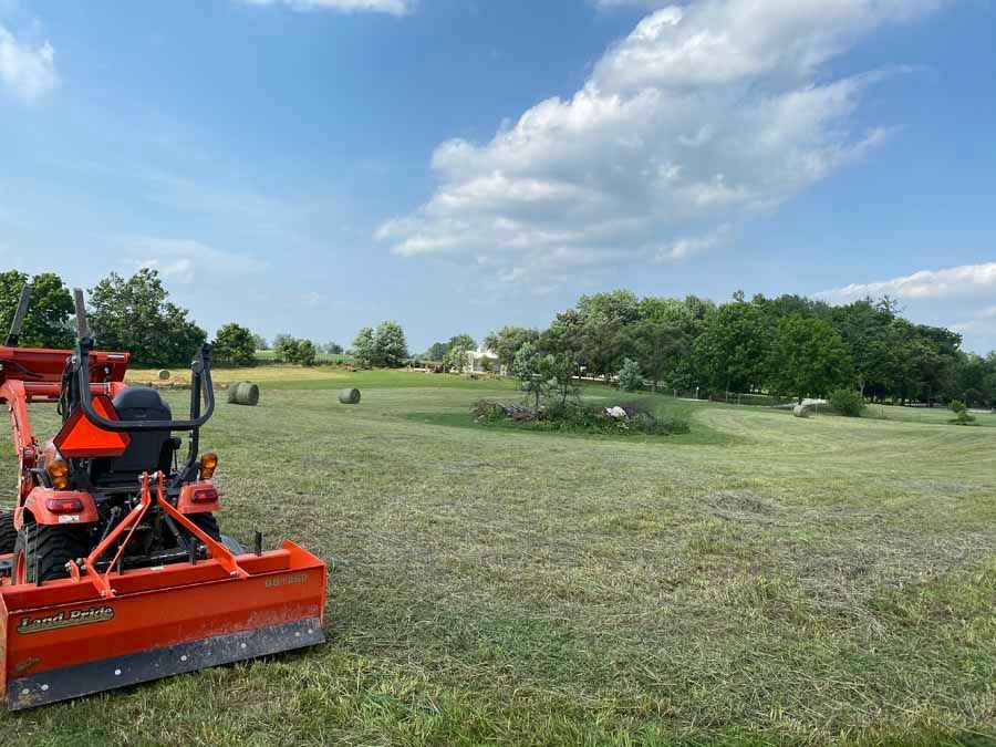 An orange tractor with a rear box blade stands in a large, mown field under a blue sky with scattered clouds.