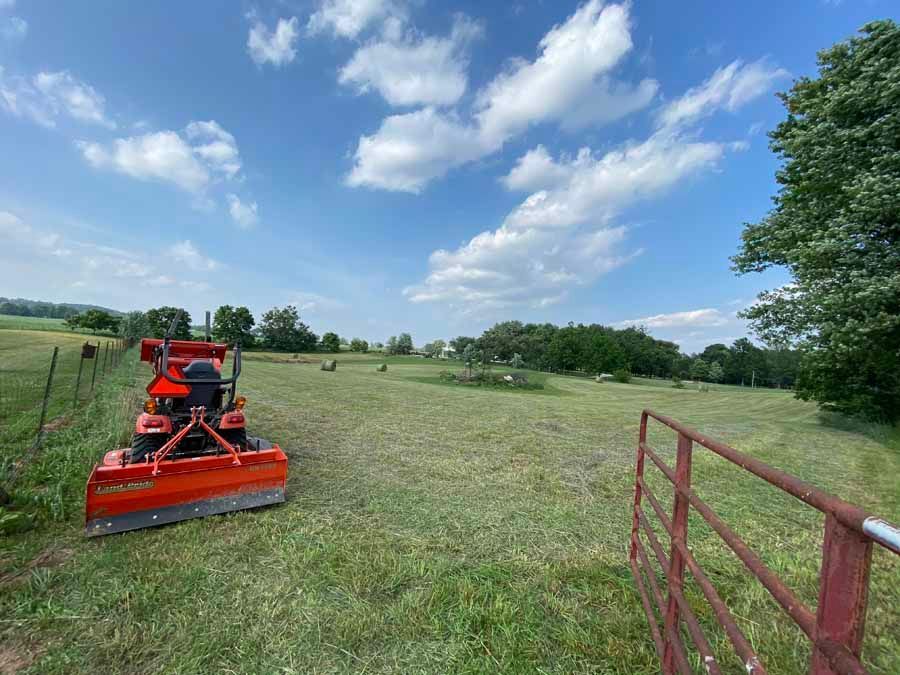 An orange tractor with a mower attachment sits in a mowed field next to a wire fence and a red metal gate under blue skies.