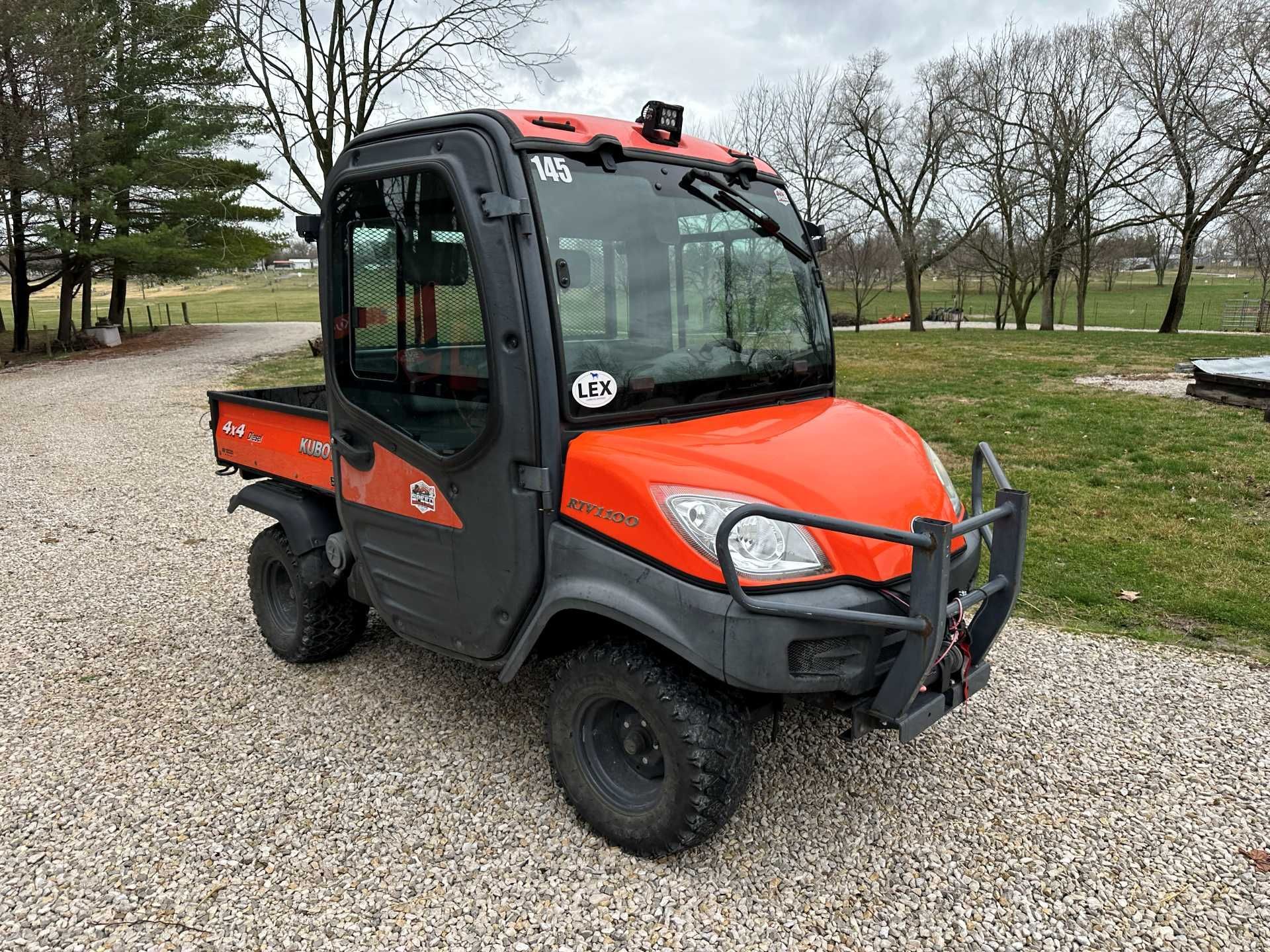 An orange and grey Kubota utility vehicle with an enclosed cab parked on a gravel path against a background of trees.