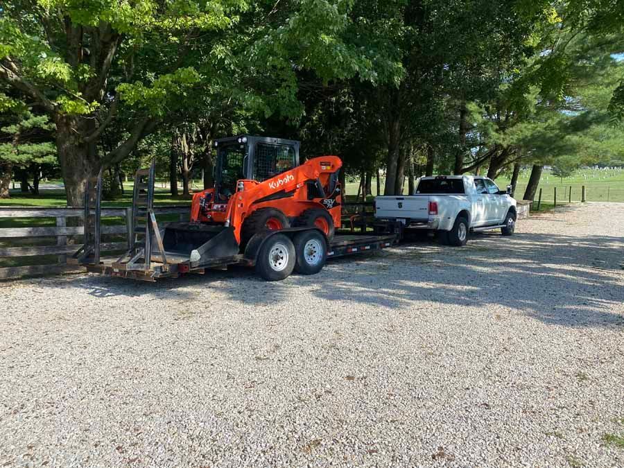 A white pickup truck towing a flatbed trailer with an orange skid-steer loader parked on a gravel surface near trees.