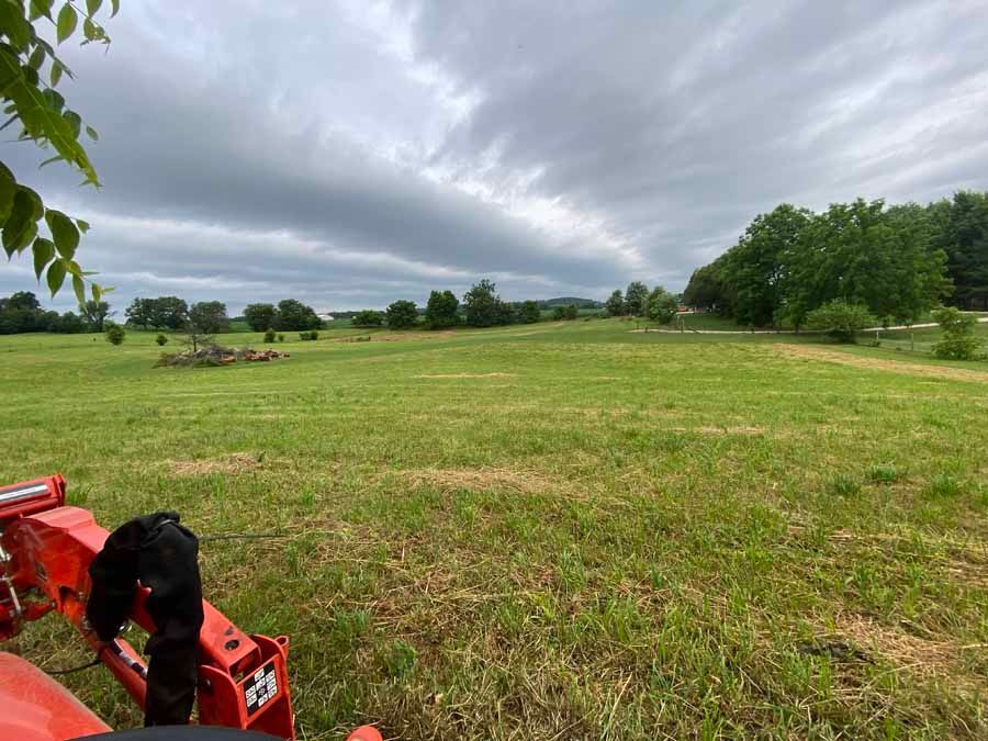 A red tractor attachment sits in the foreground of a large, grassy field under a cloudy, overcast sky.
