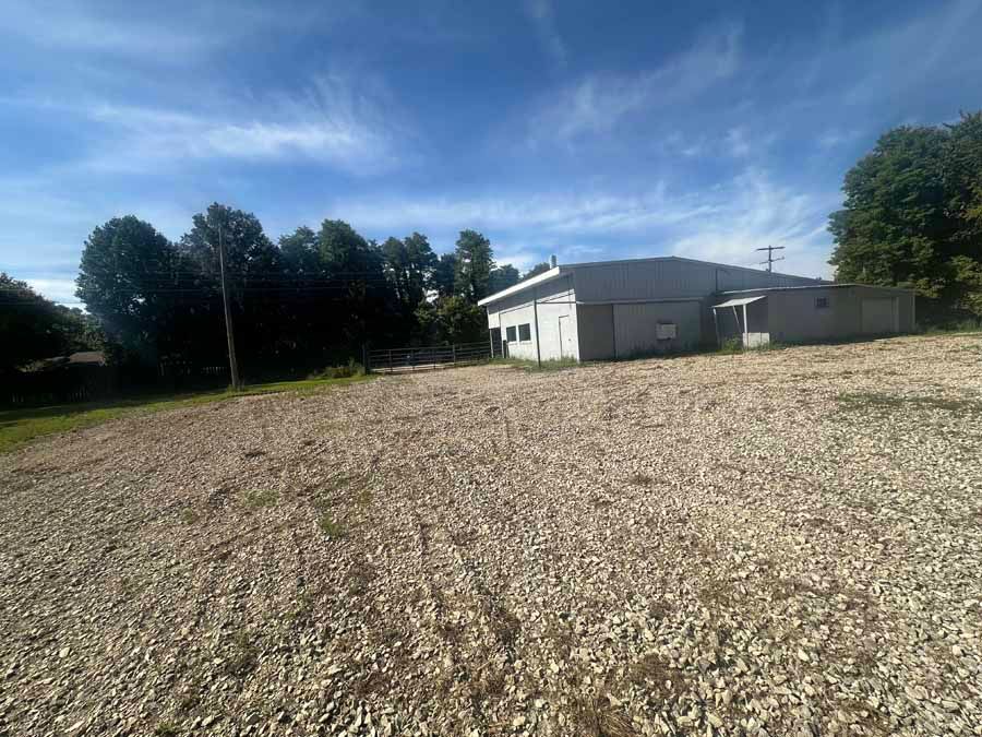 A gravel-covered lot leading to a long, light-gray industrial building framed by a line of trees under a blue sky.