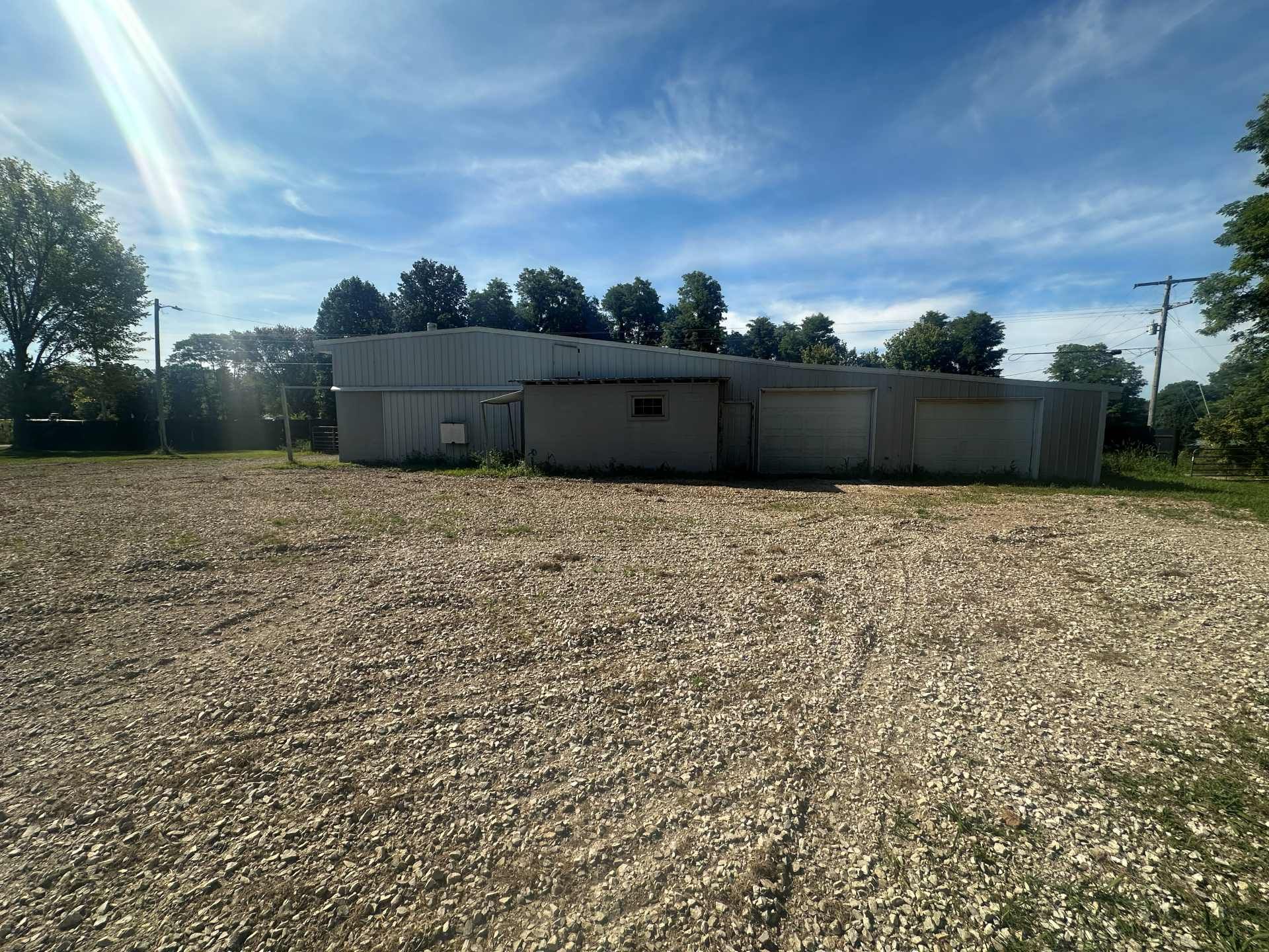 A long, low-profile industrial building sits behind a wide, dry field covered in light-colored gravel under a blue sky.