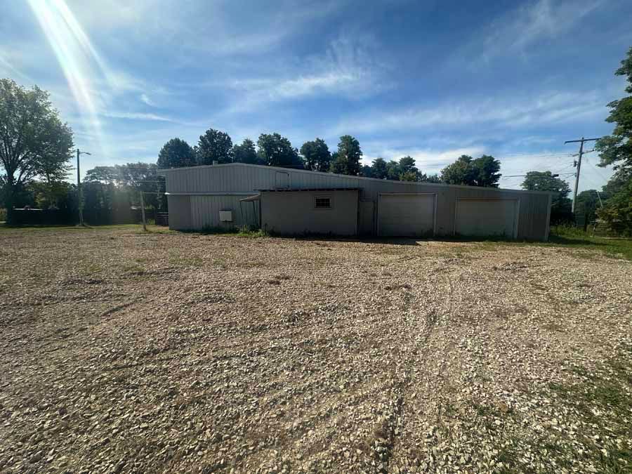 A long, gray metal warehouse building sits in the middle of a large, gravel-covered lot under a bright, sunny sky.