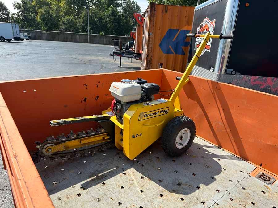A yellow gas-powered walk-behind trencher sits inside the metal bed of a flatbed trailer.
