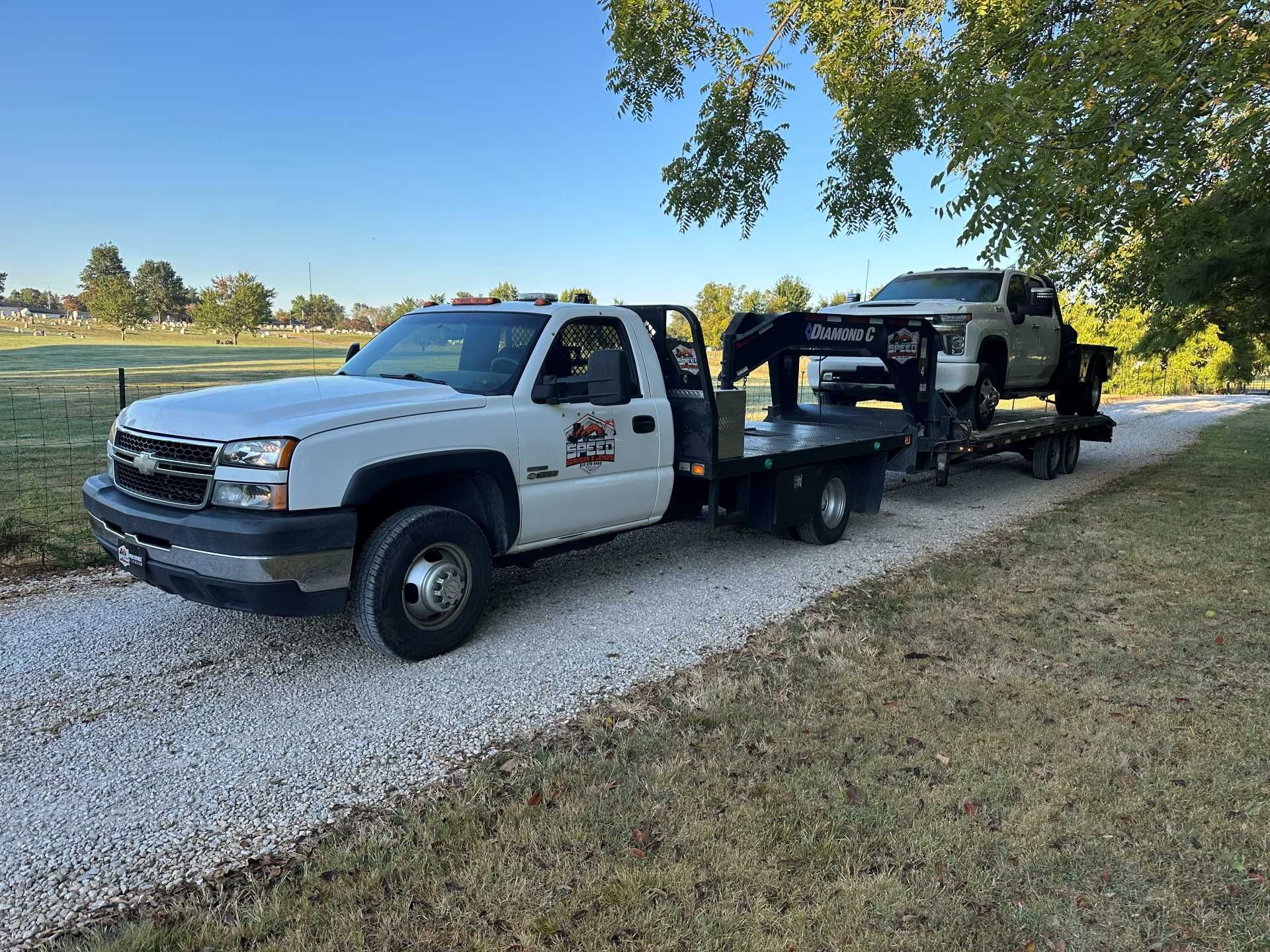 A white flatbed tow truck hauls a white pickup truck on a gooseneck trailer along a gravel road near trees.