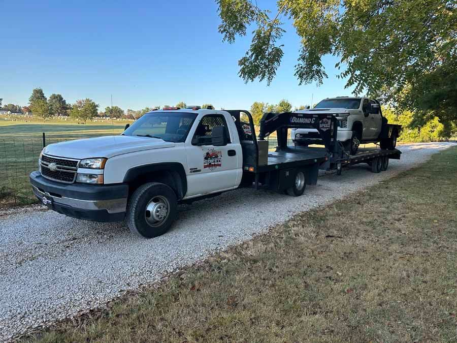 A white pickup truck with a flatbed trailer hauling a white SUV on a gravel driveway under a clear blue sky.