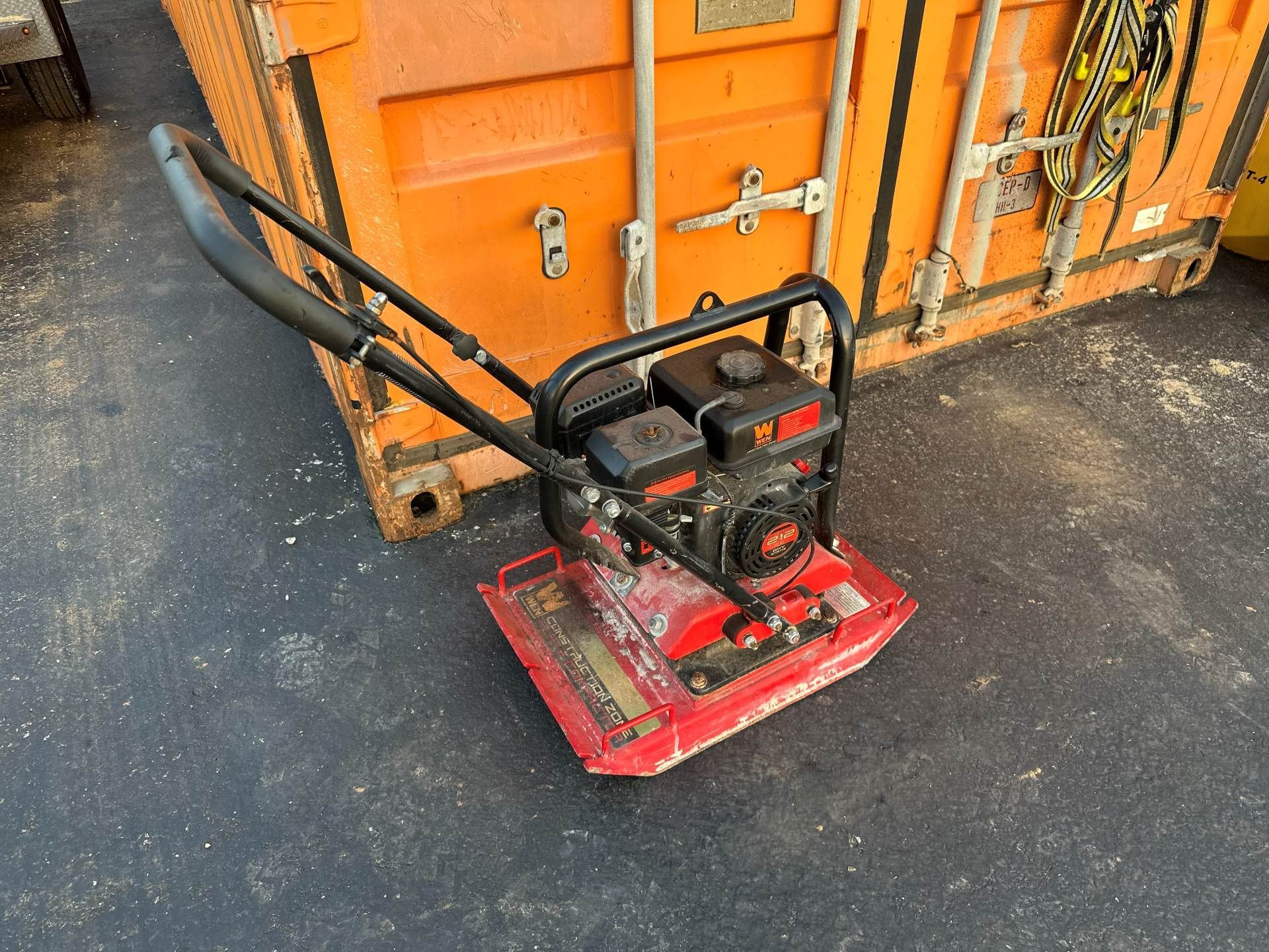 A red gas-powered plate compactor sitting on asphalt next to an orange shipping container.