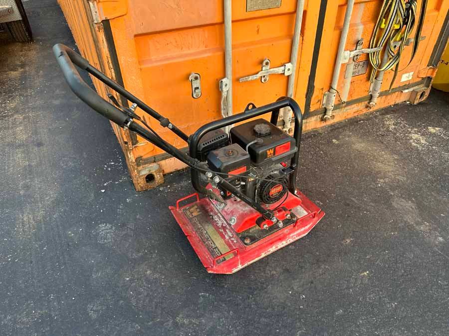 A red plate compactor sits on a dark paved surface in front of an orange storage container.