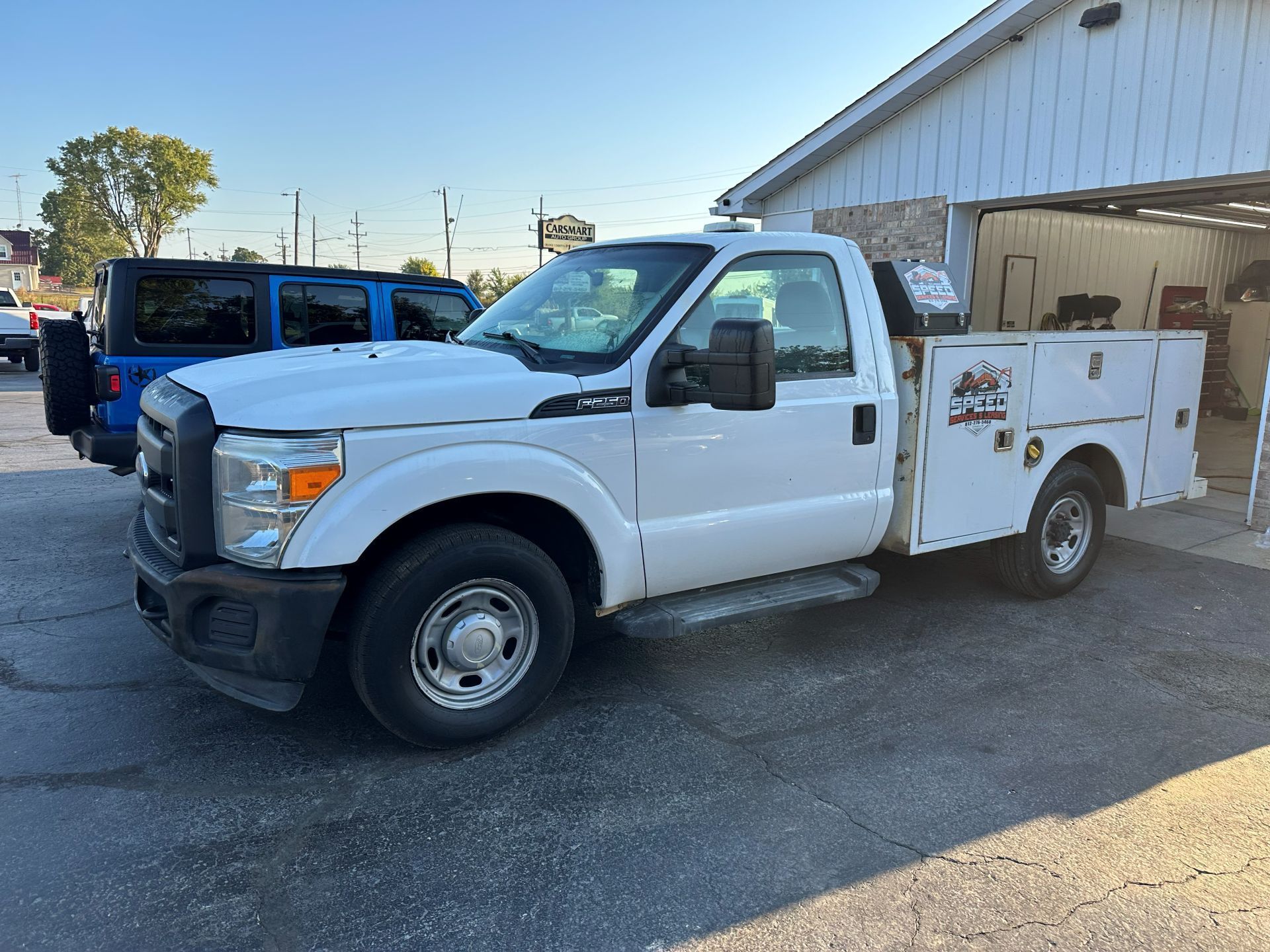 A white Ford F-series service truck parked on asphalt next to a blue SUV outside a white industrial building.