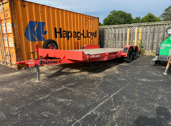 A red flatbed utility trailer parked on an asphalt lot next to a large orange Hapag-Lloyd shipping container.