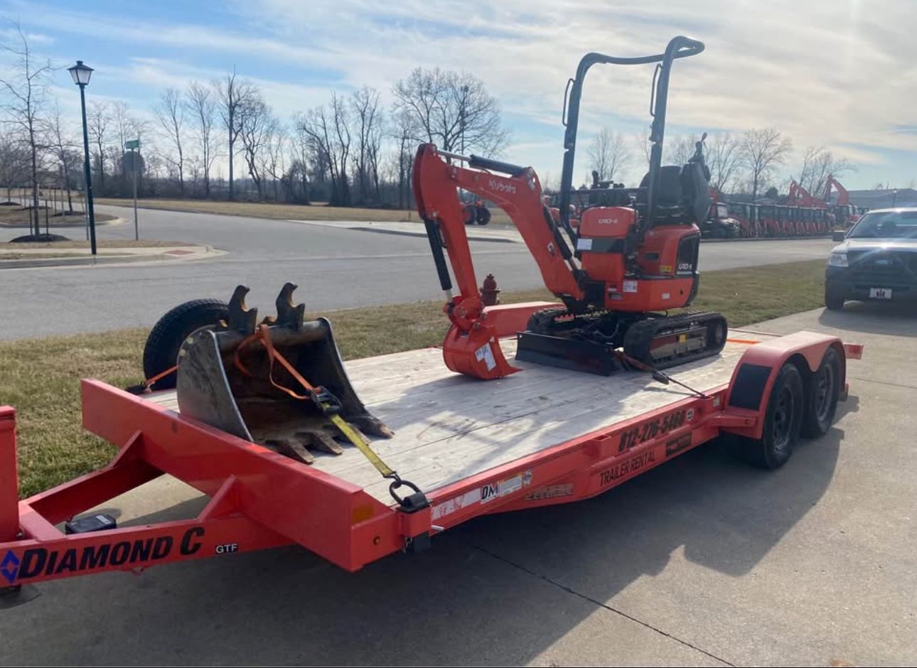 A bright orange Kubota mini excavator with a spare bucket is secured to a Diamond C utility trailer on an asphalt lot.