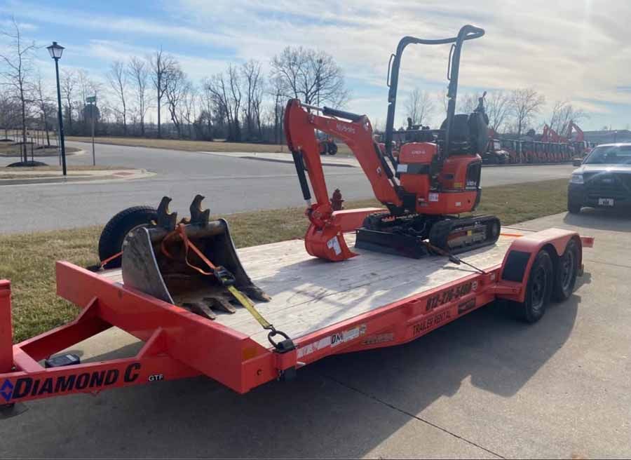 A bright orange Diamond C flatbed trailer carrying a compact orange excavator and a large metal bucket, parked outdoors.