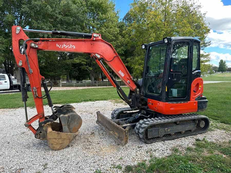 A bright orange Kubota compact excavator with a closed cab and rubber tracks parked on a gravel lot.