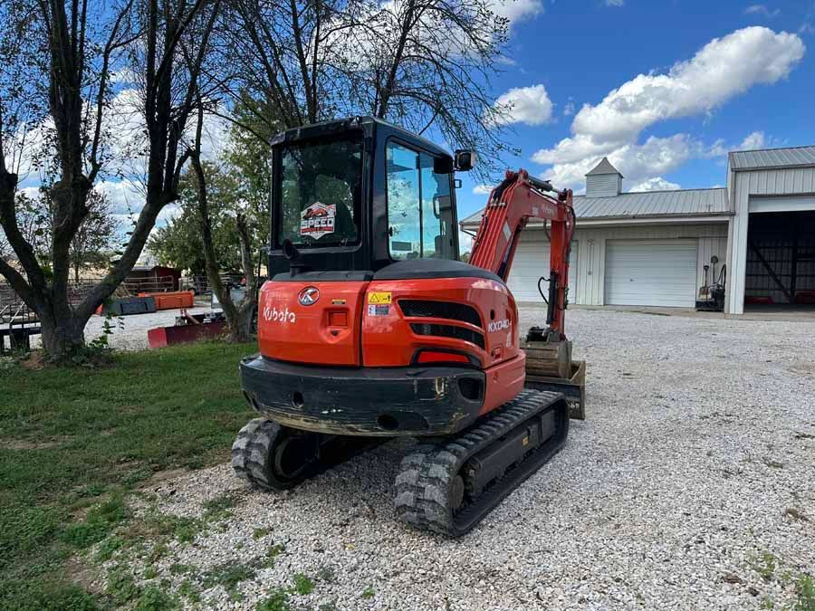 An orange Kubota compact excavator parked on a gravel lot in front of a white building under a sunny, partly cloudy sky.