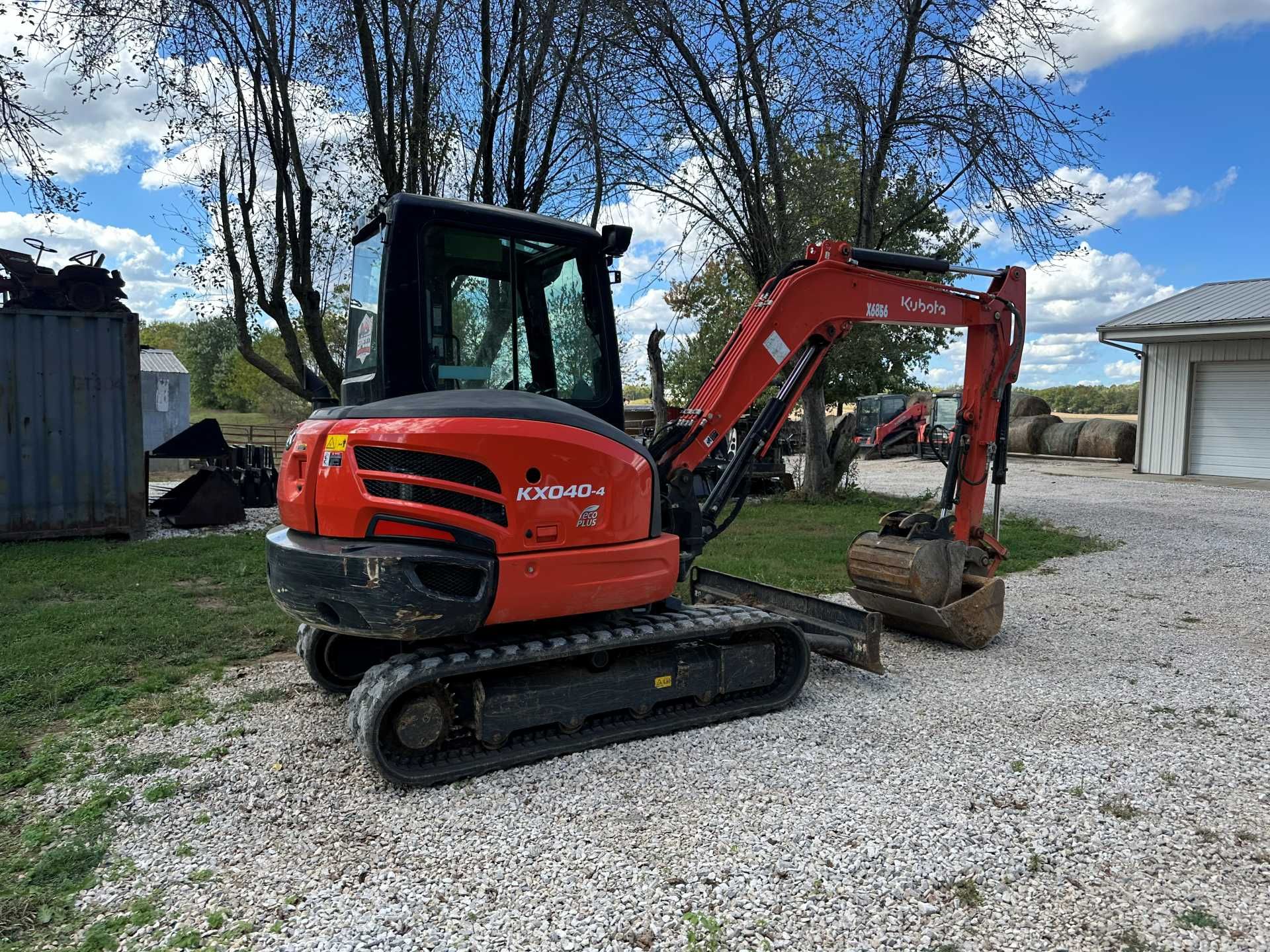 An orange Kubota compact excavator parked on a gravel surface with trees and a building in the background.