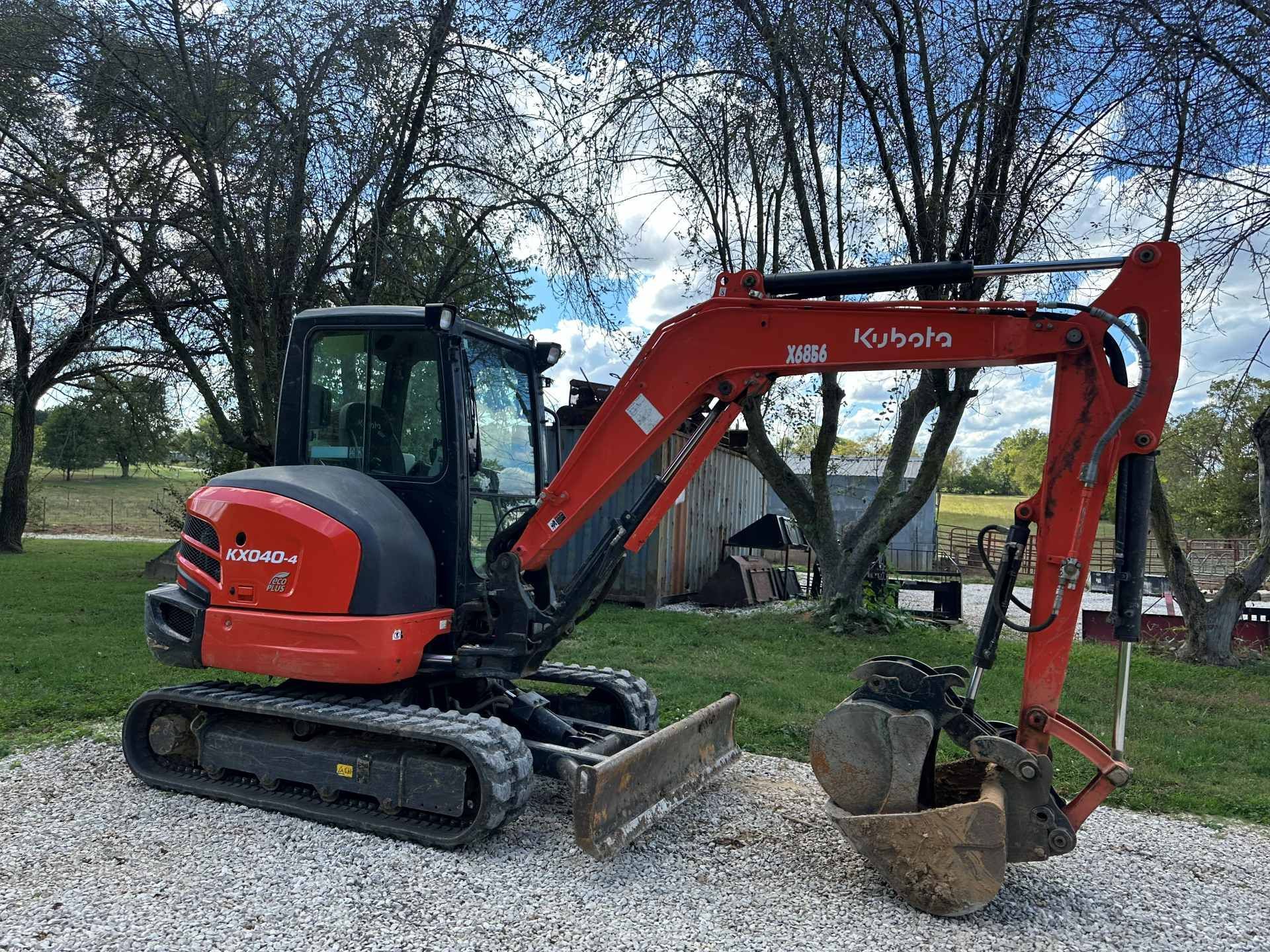 A bright orange Kubota compact excavator parked on a gravel surface outdoors with trees in the background.