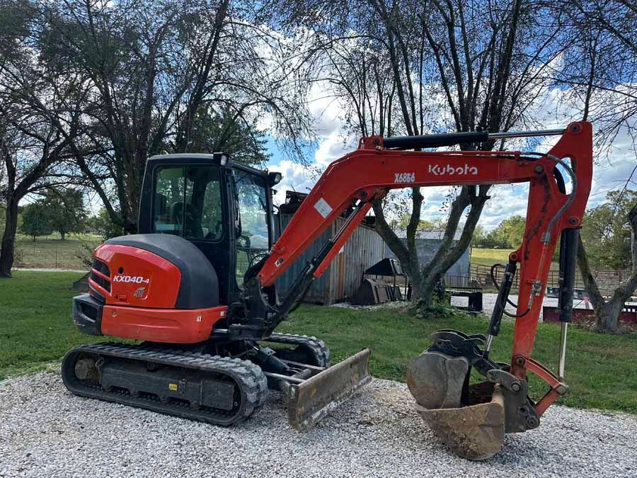 An orange Kubota compact excavator parked on a gravel surface outdoors with a grassy field and trees in the background.