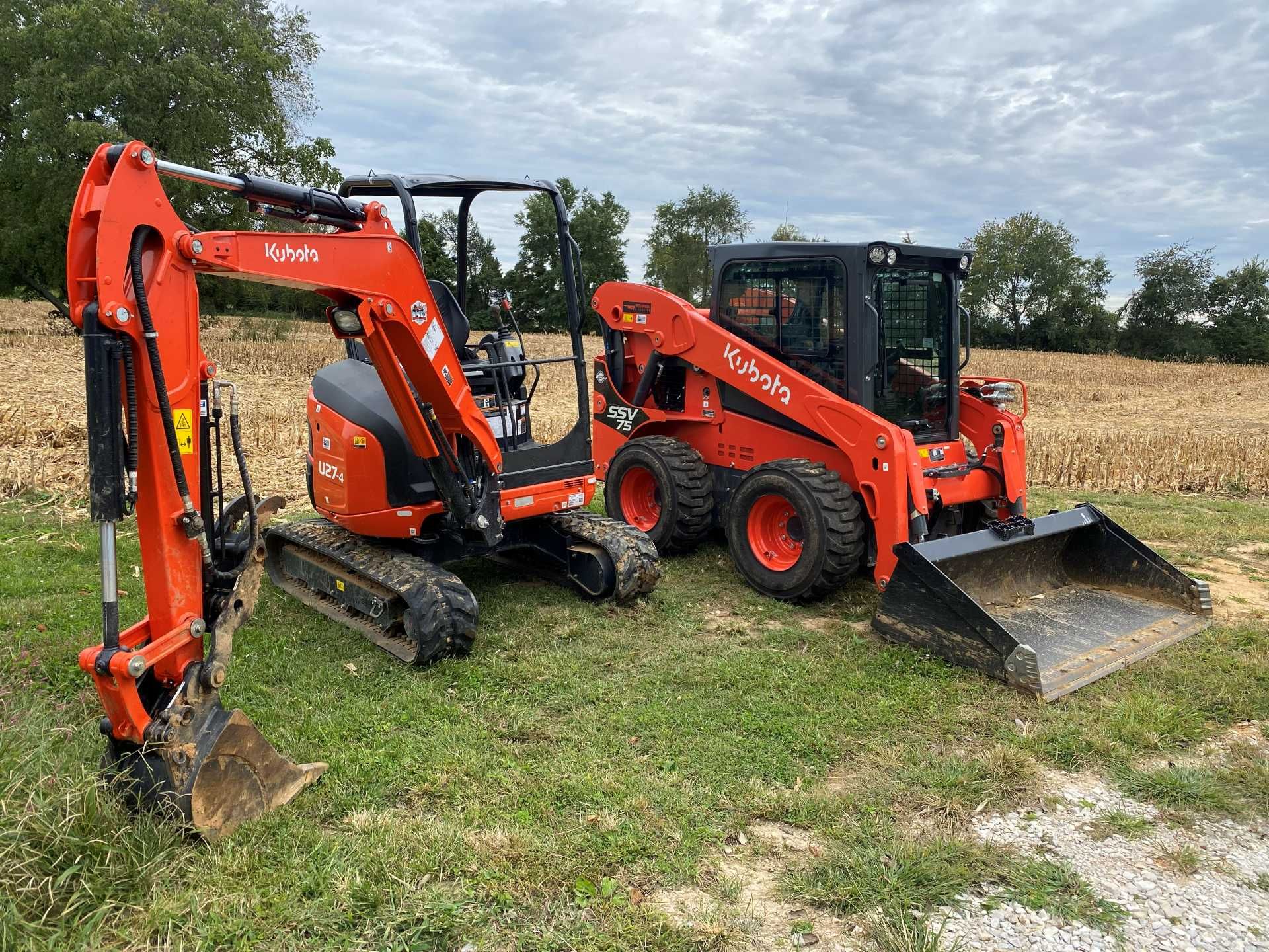 Two orange Kubota construction machines, an excavator and a skid steer, parked in a grassy field.