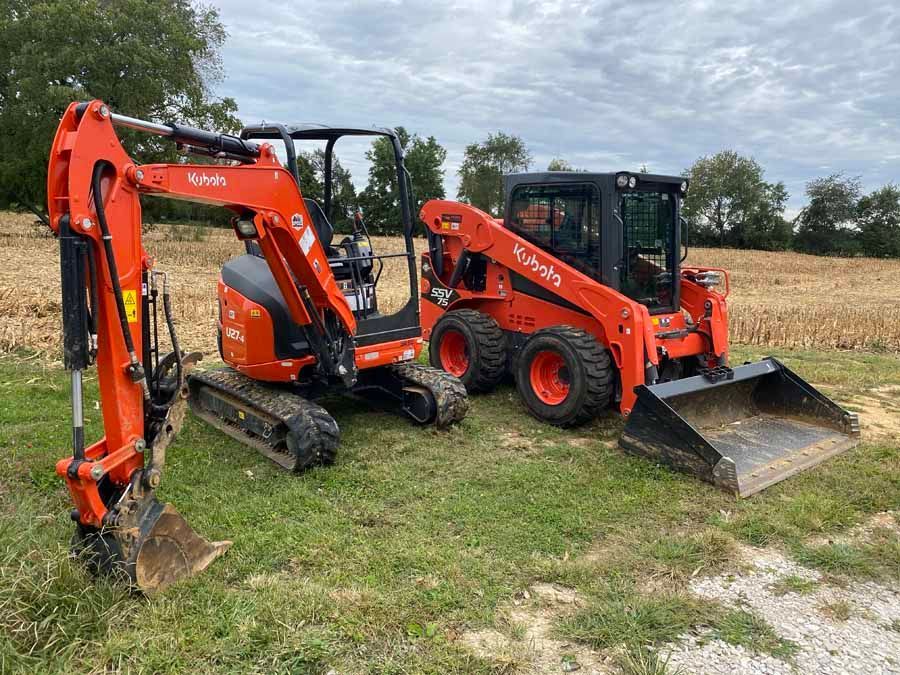 An orange Kubota excavator and a matching skid steer parked in a field with a treeline and cloudy sky in the background.