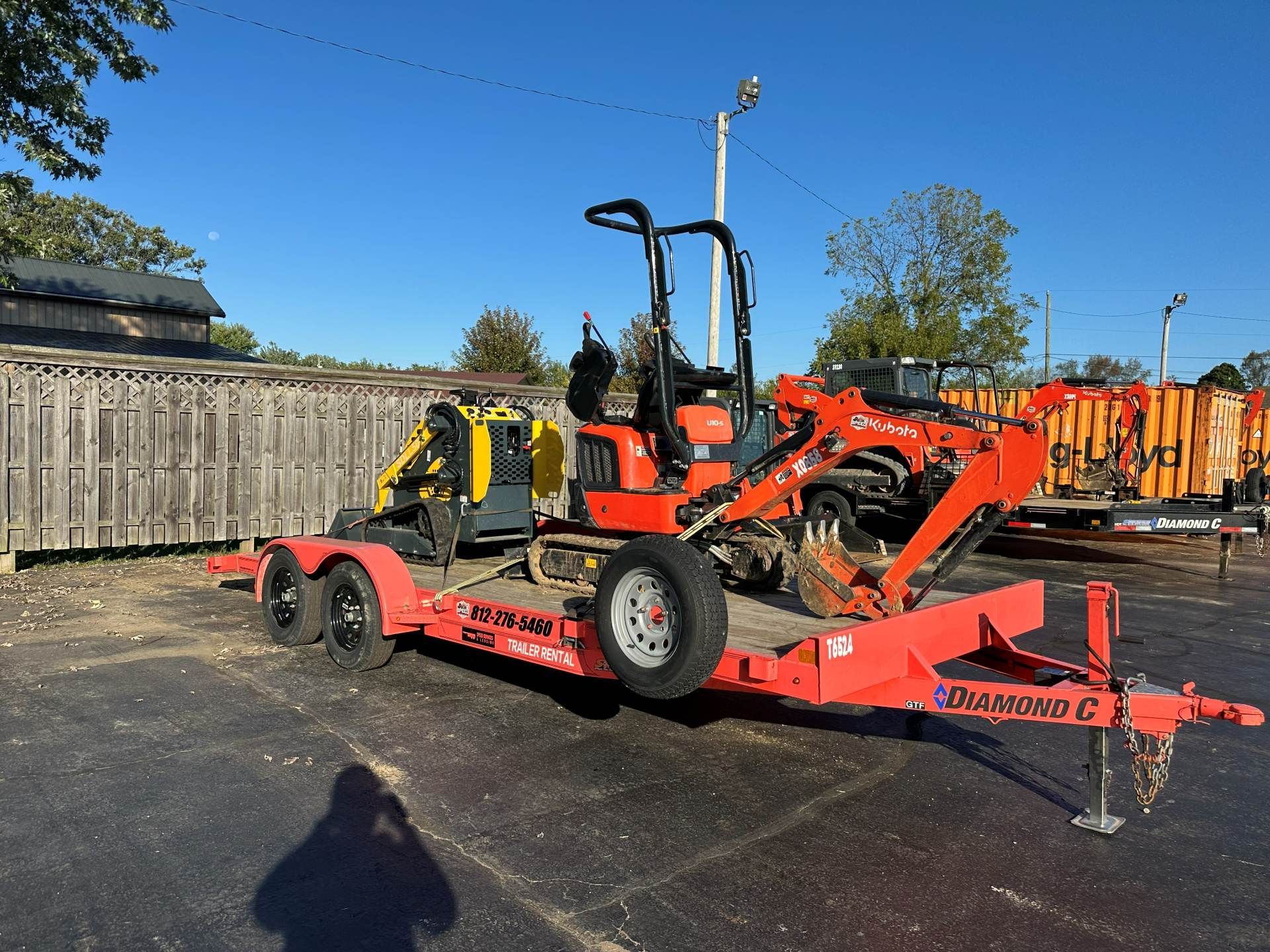 An orange flatbed trailer carrying a small orange excavator and a yellow skid steer loader parked on a paved lot.