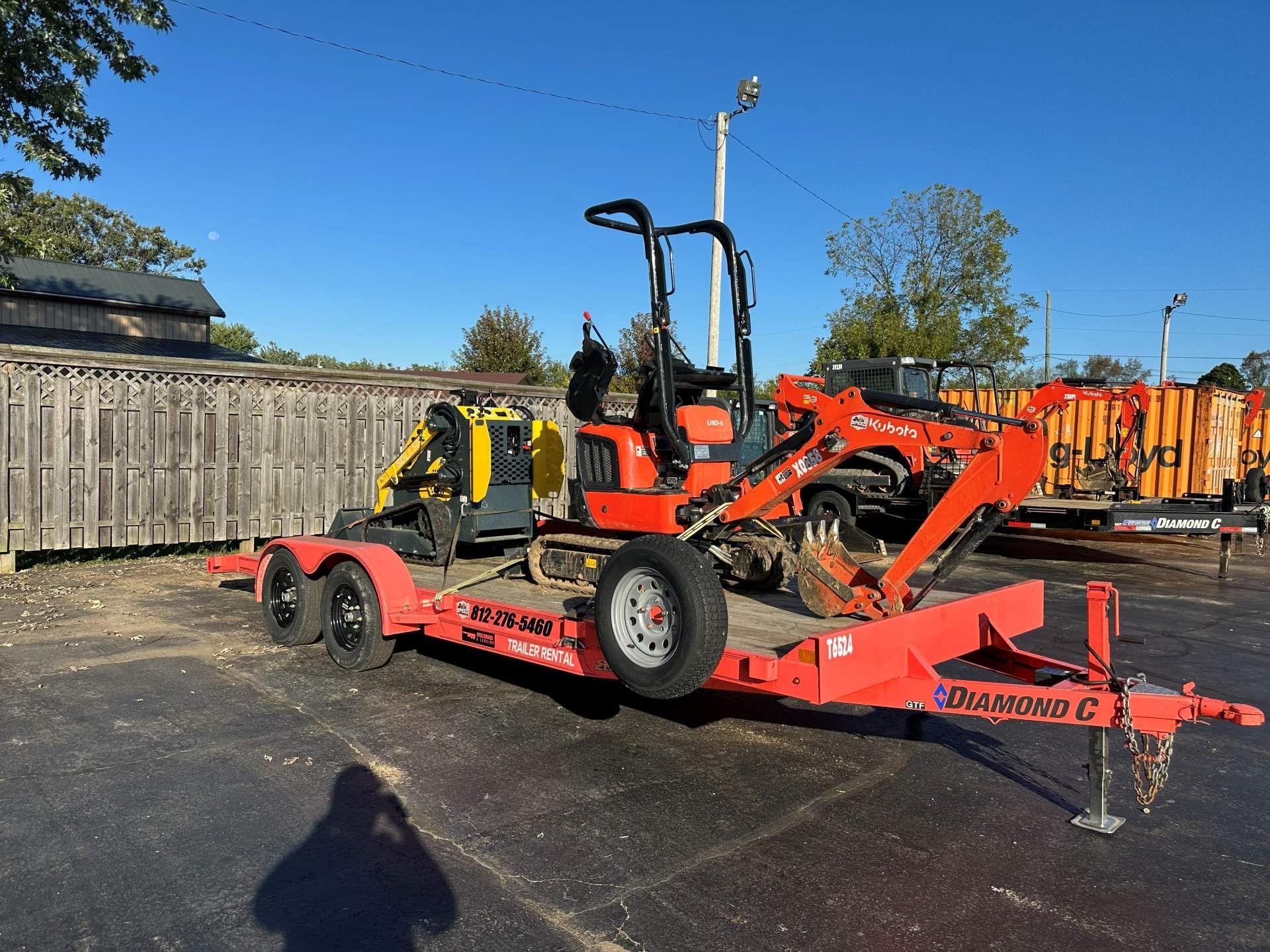 A bright orange flatbed trailer parked on asphalt, carrying a yellow skid steer loader and an orange mini excavator.