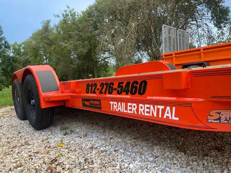 An orange rental trailer with the phone number 812-276-5460 printed on the side, parked on a gravel surface.