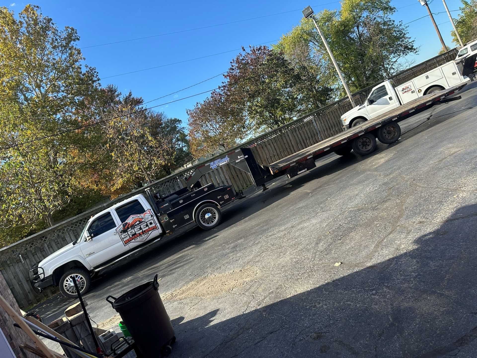 A white flatbed truck parked in a gravel lot with a second white vehicle loaded onto its trailer.