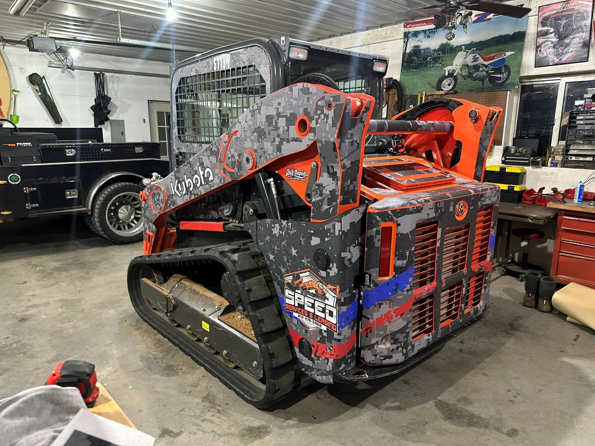 A Kubota compact track loader with a custom grey camouflage and orange paint job parked inside a workshop.