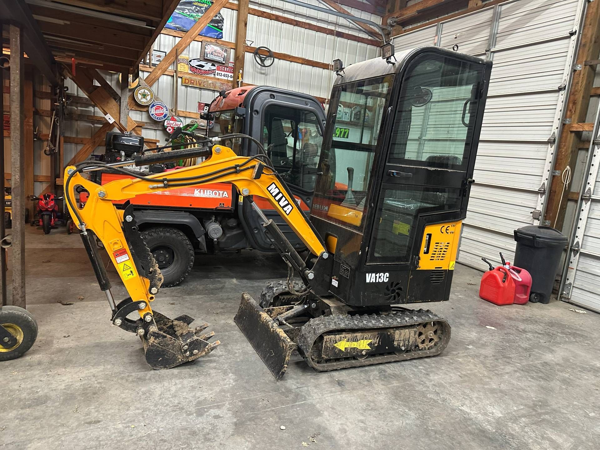 A yellow and black mini excavator parked inside a shed, partially blocking an orange utility vehicle in the background.