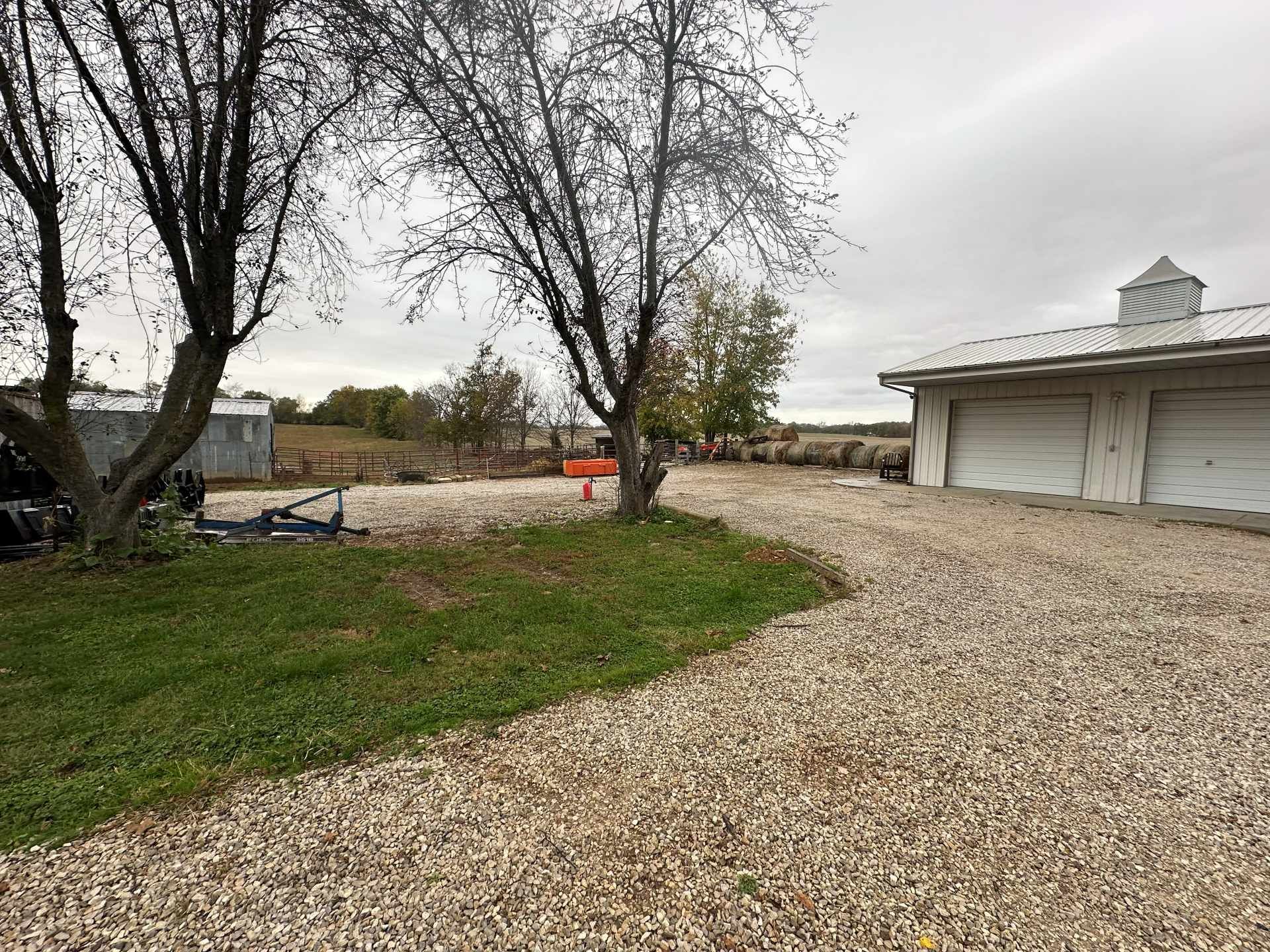 A gravel driveway leads to a light-colored multi-car garage on a cloudy day, framed by two large trees in the foreground.