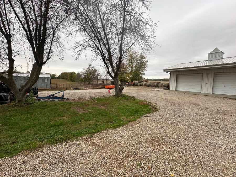 A gravel driveway leads toward a light-colored barn and a secondary shed under a cloudy sky.