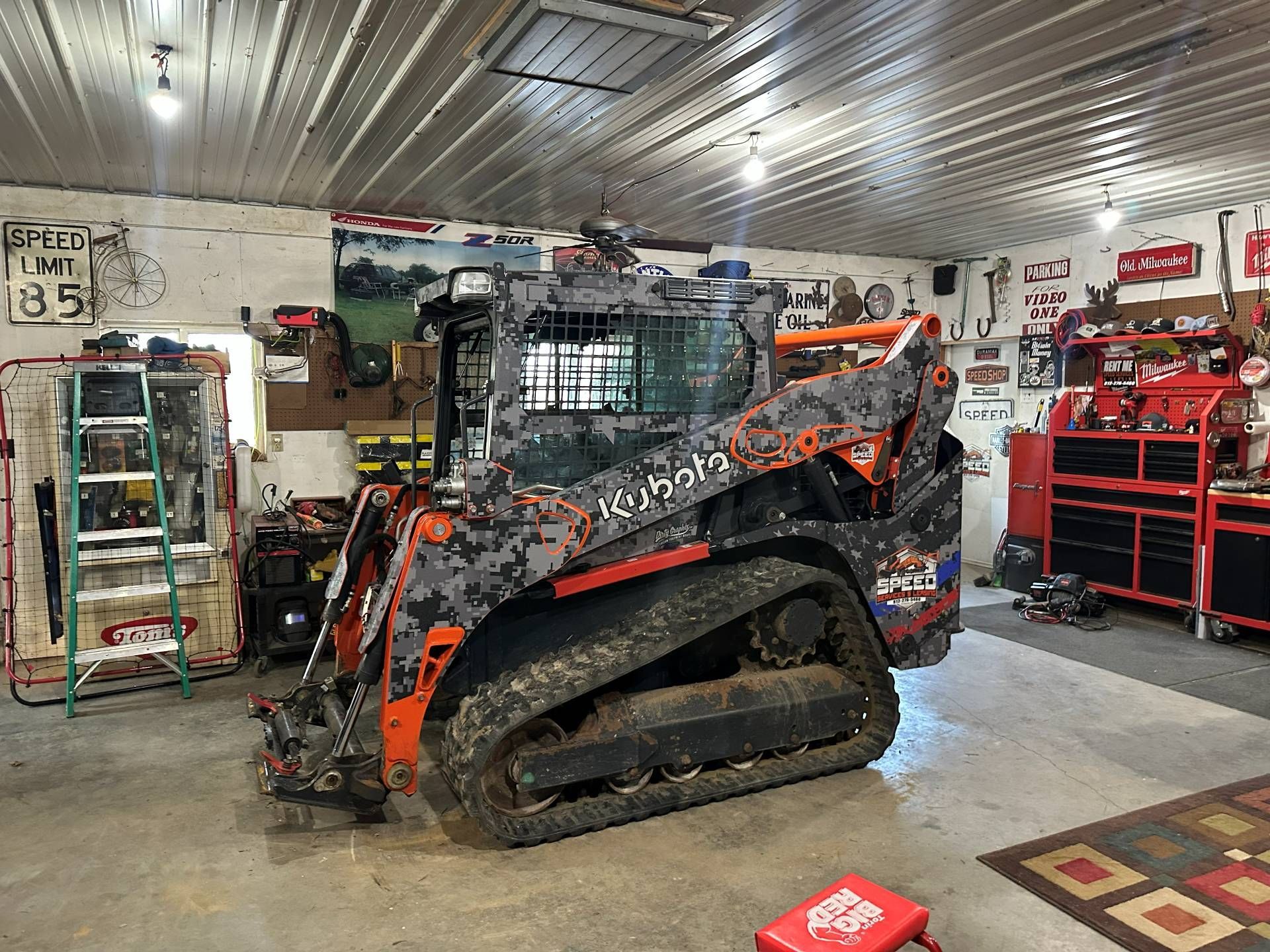 A grey camo-patterned Kubota compact track loader parked in a well-lit workshop with tools and cabinets in the background.