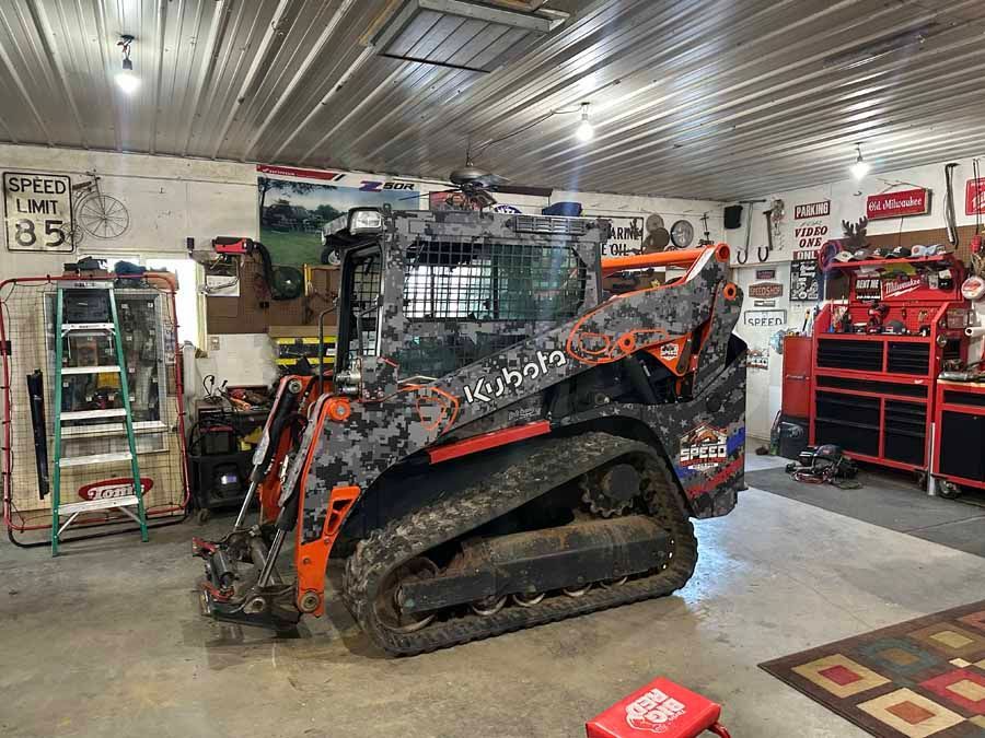 A custom camouflage-patterned Kubota skid steer loader parked inside a well-equipped, cluttered garage workshop.