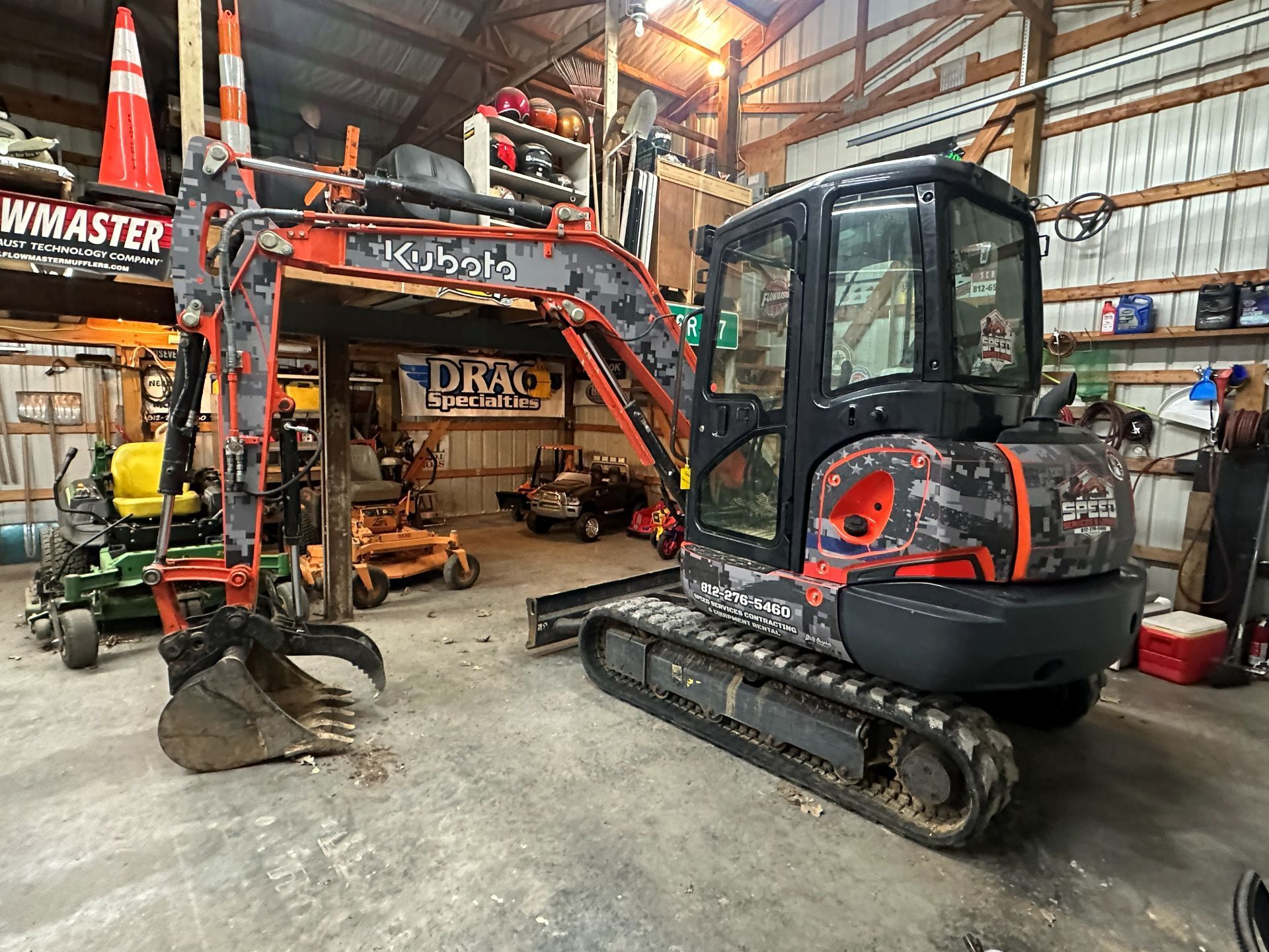 A camouflage-patterned Kubota mini excavator with a bucket attachment parked inside a cluttered workshop or garage.