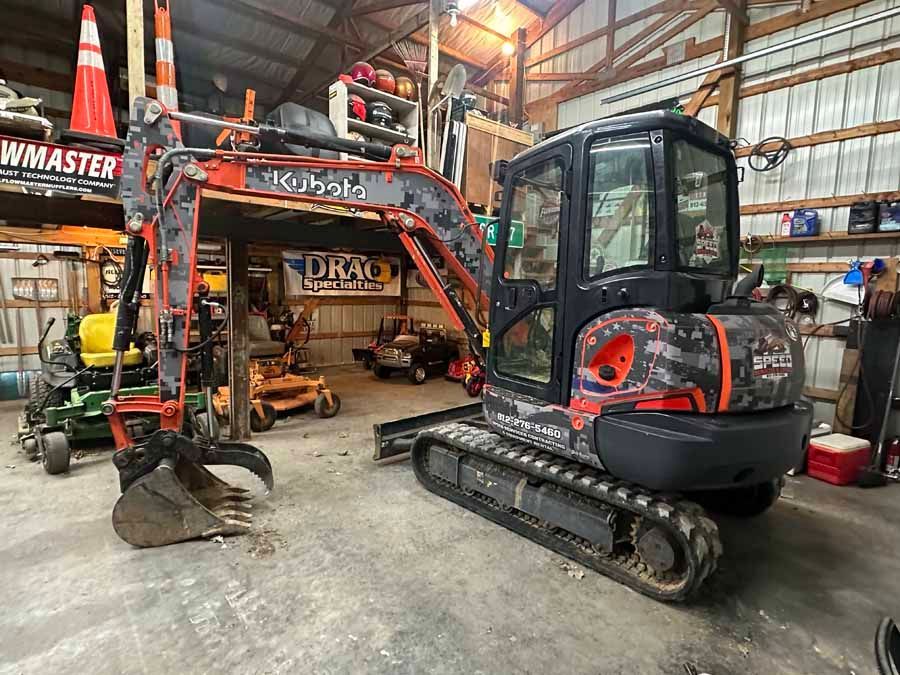 A Kubota mini excavator with camouflage body panels parked inside a cluttered equipment workshop.
