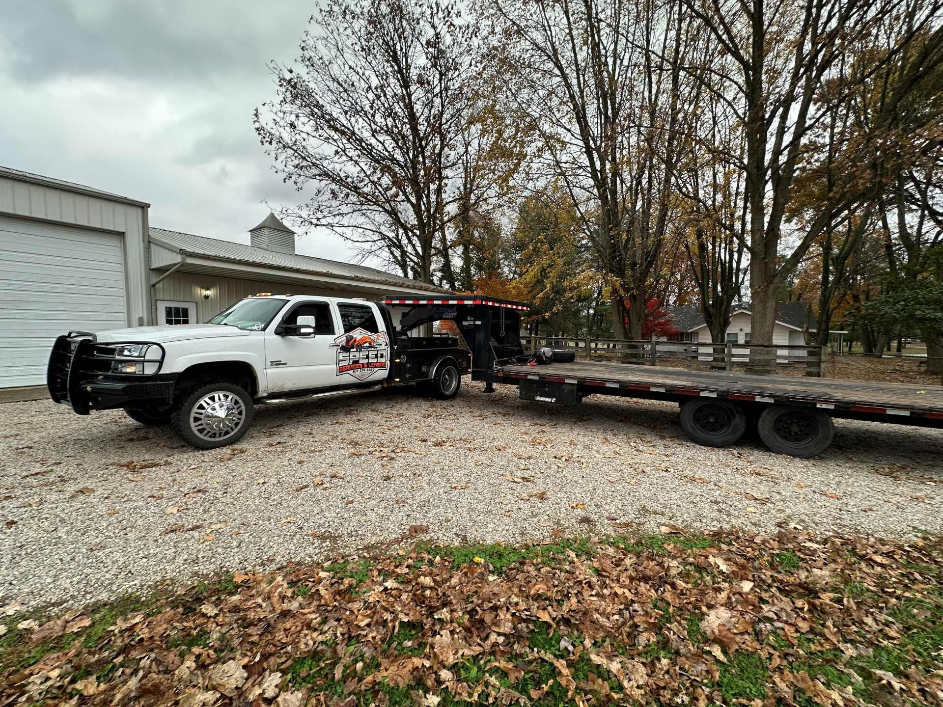 A white flatbed truck with a large bull bar and logo is hitched to an empty flatbed trailer in a gravel lot.