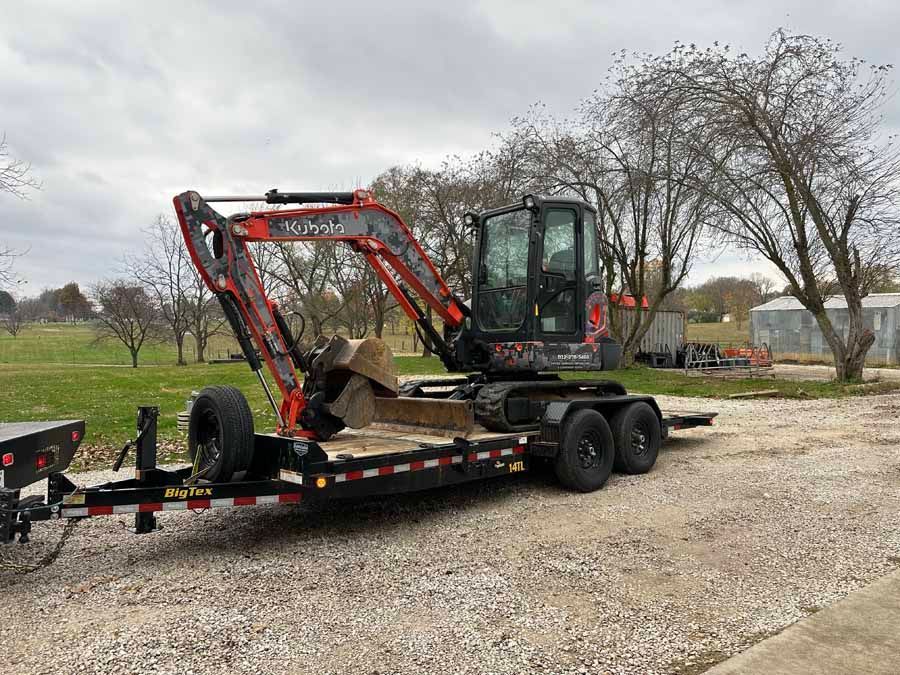 An orange and black Kubota compact excavator loaded on a flatbed trailer parked on a gravel lot under a cloudy sky.