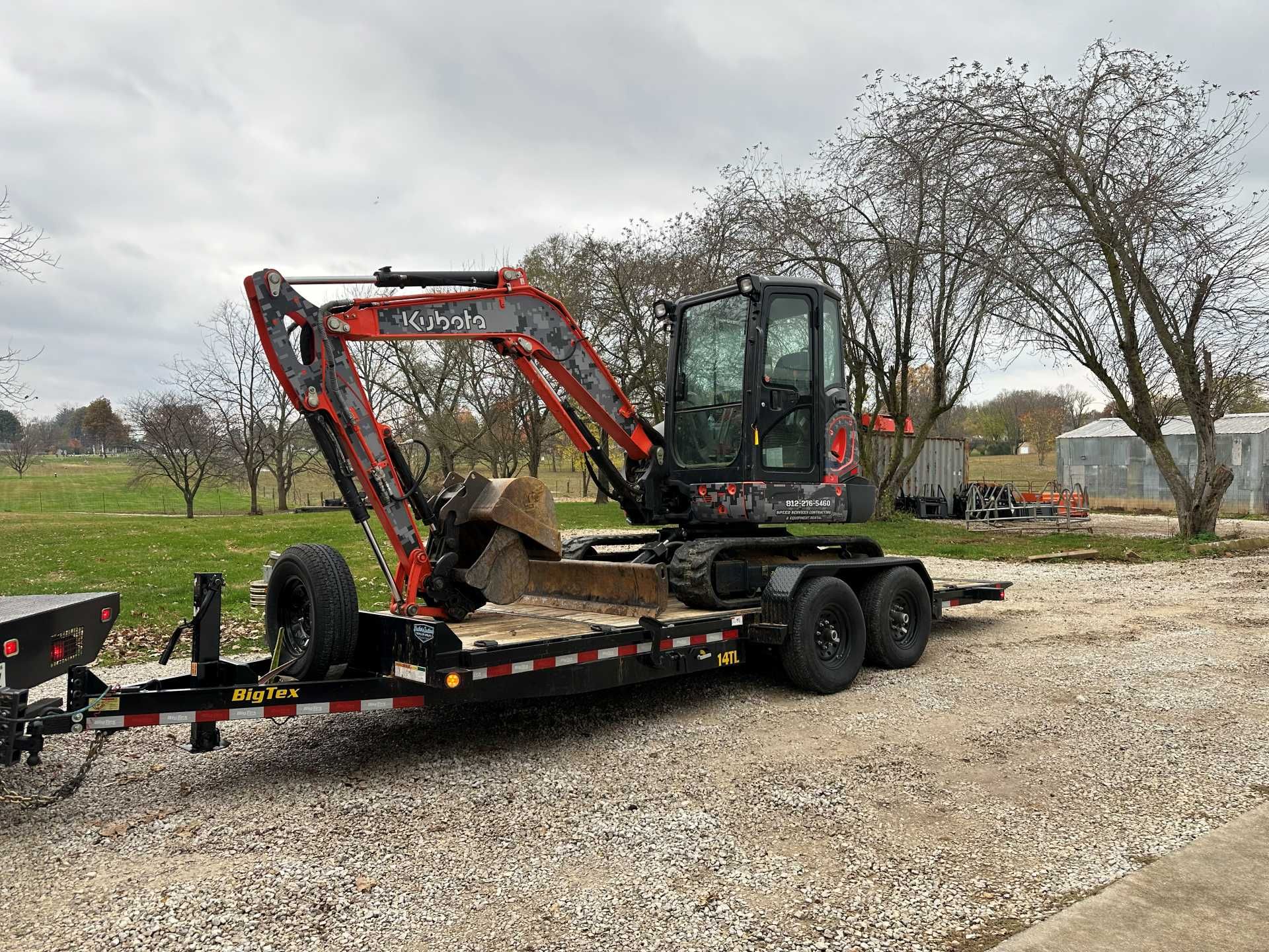 An orange Kubota excavator sits secured on a black flatbed trailer parked on a gravel lot under a cloudy sky.
