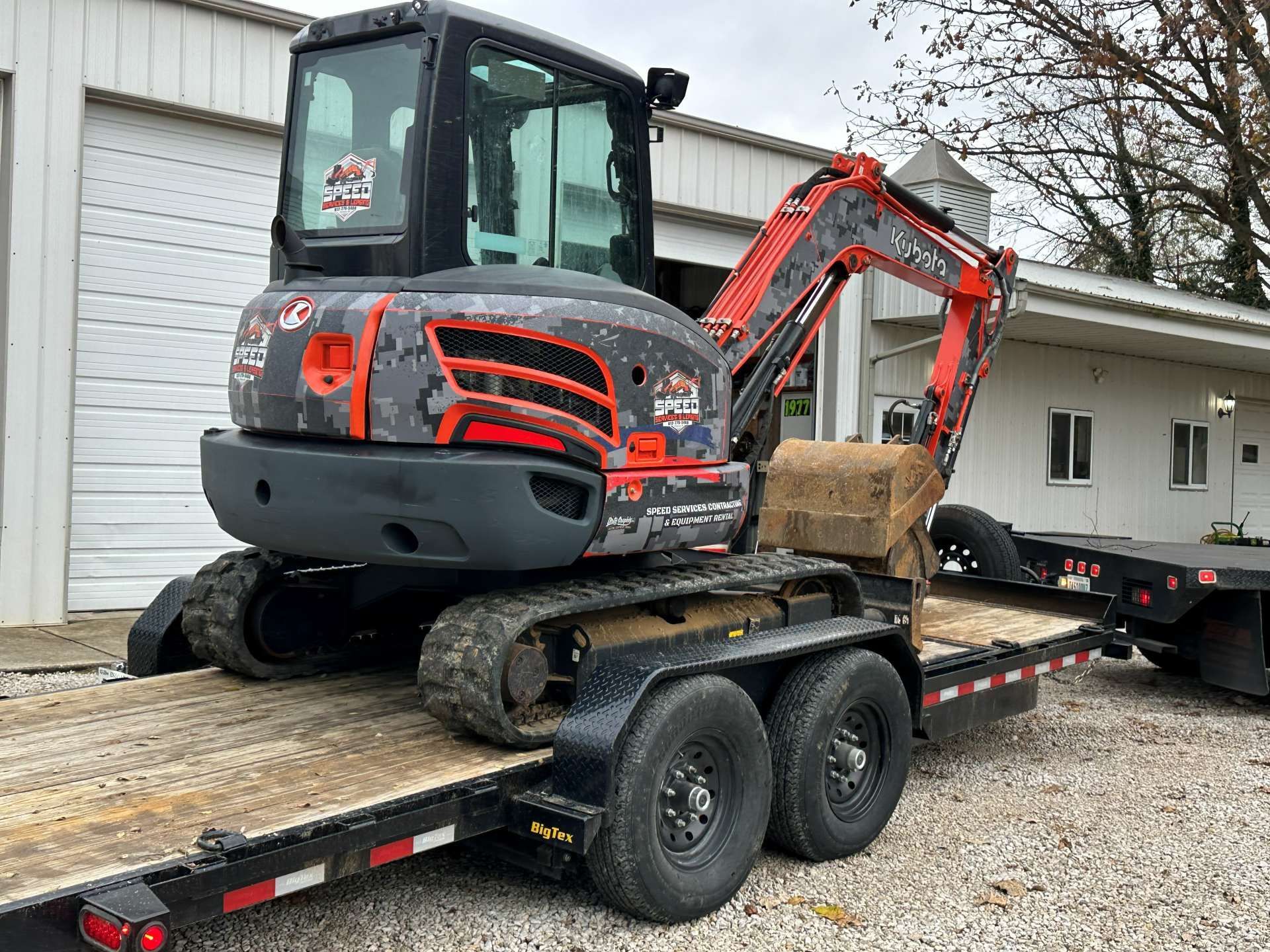 A gray and orange Kubota mini excavator loaded onto a flatbed trailer parked outside a white industrial building.
