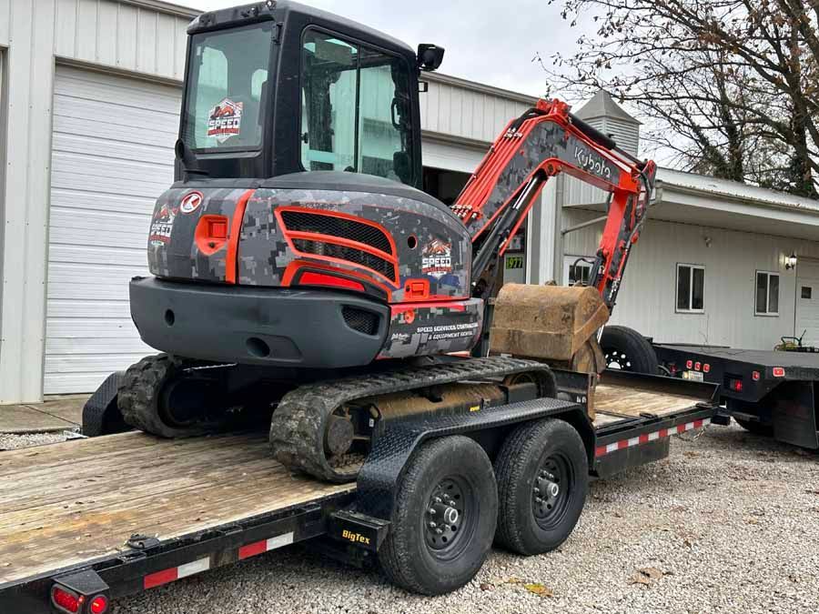 A Kubota excavator with a gray camouflage wrap sits parked on a flatbed trailer outdoors in front of a building.