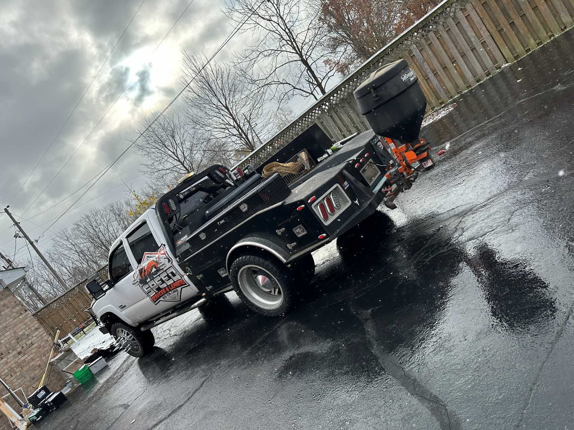 White and black work truck with a salt spreader attached, parked on wet asphalt in front of a fence.