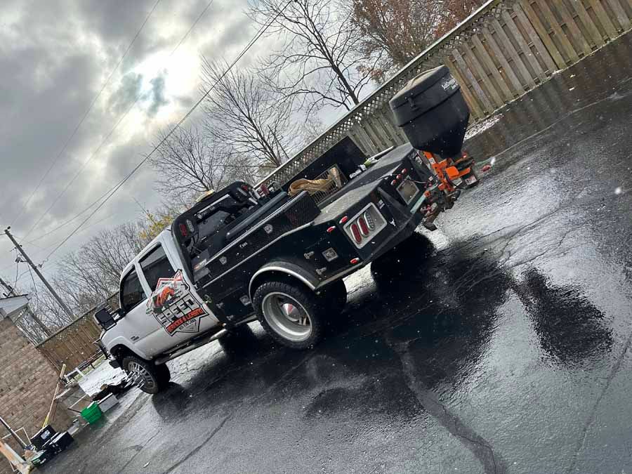 A white pickup truck with a black flatbed utility body and salt spreader parked on wet pavement under a cloudy sky.